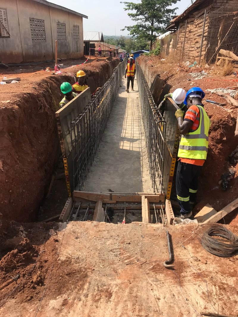 Construction workers in hard hats and safety vests building a reinforced concrete drainage channel in a deep excavation trench, with rebar cages and timber formwork visible.
